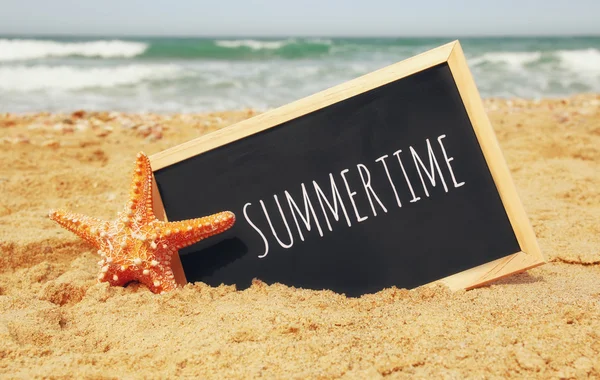 Starfish e clachboard com frase: SUMMERTIME, na areia do mar e horizonte oceânico — Fotografia de Stock