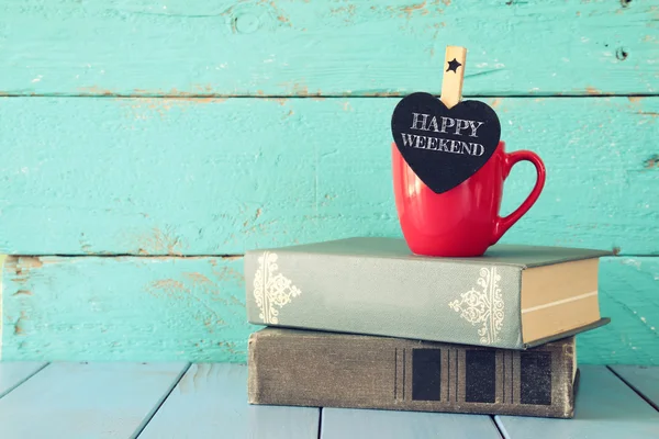 Tasse Kaffee mit einer kleinen herzförmigen Tafel mit der Aufschrift: Frohes Wochenende und zum Stapeln alter Bücher. — Stockfoto