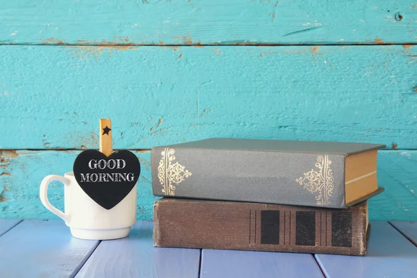 Cup of coffee with a little heart shape chalkboard with the phrase: GOOD MORNING next to stack of old books. — Stock Photo, Image