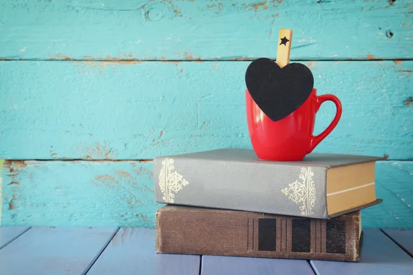 Чашка кофе с маленькой доской в форме сердца и стопка старых книг . — стоковое фото