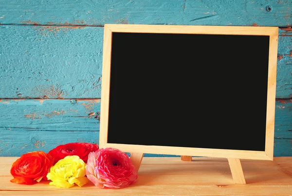 Flowers next to empty blackboard, on wooden table. copy space. — Stock Photo, Image