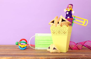 Ahşap masa ve mor arka plan üzerine Purim bayramı konsepti (Musevi karnavalı). Coronavirus önleme konsepti, tıbbi maske