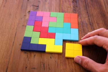 Bir adamın eli ahşap bir masanın üzerinde kare bir tangram yapbozu tutuyor.