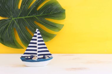 Beyaz dekoratif yelkenli tekne, ahşap masa üzerinde deniz kabukları ve sarı arka plan