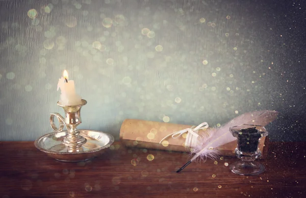 Низкое ключевое изображение белого перышка, чернильницы, свитка и свечи и блестки светильников фона на старом деревянном столе — стоковое фото