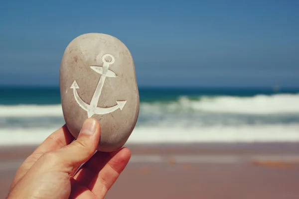 Man's hand holding one stone pebbles with theanchor sign  against sandy beach and sea horizone — Stock Photo, Image