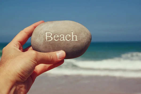Mans hand håller en sten stenar med theanchor skylt mot sandiga stranden och havet horizone — Stockfoto