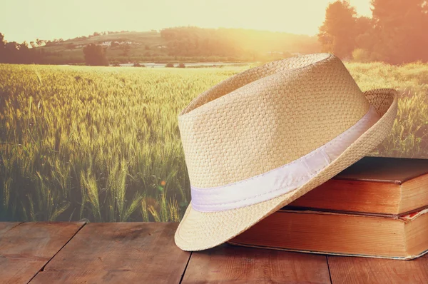 Fedora hoed en stapel boeken over houten tafel en gebied van tarwe land kant achtergrond. ontspanning of vakantie concept — Stockfoto