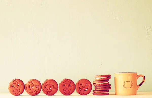 Raw of cookies with smiley face over wooden table next to cup of coffee. image is retro style filtered — Stock Photo, Image