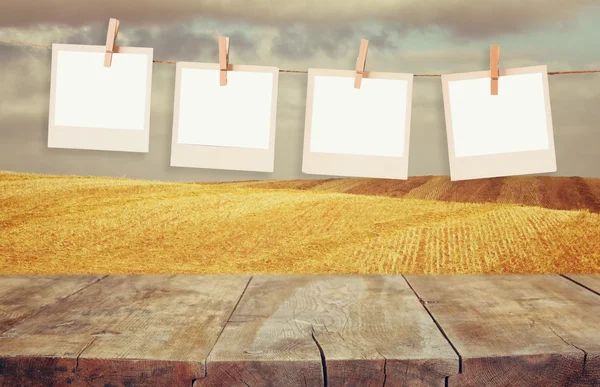 Old polaroid photo frames hanging on a rope with vintage wooden board table in front of wheat field landscape — Stock Photo, Image