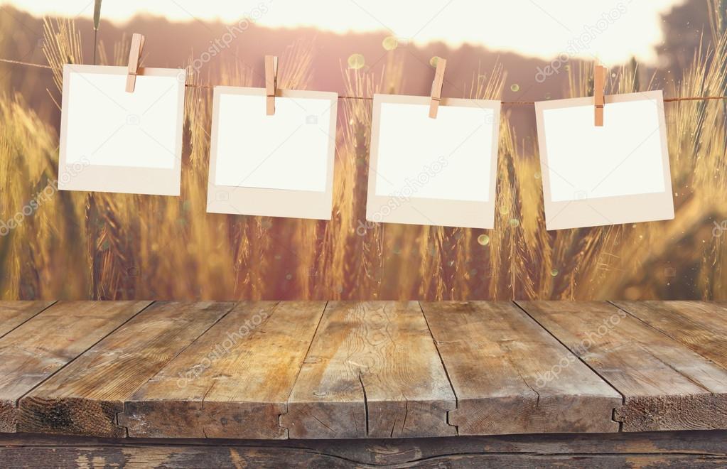 Old polaroid photo frames hanging on a rope with vintage wooden board table in front of wheat field landscape with lens flare