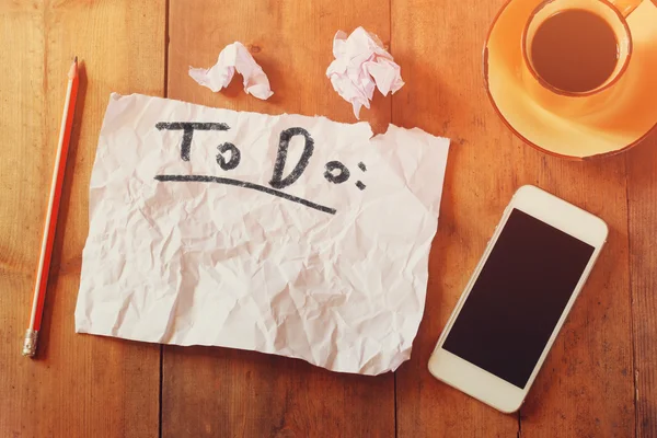 Vista superior de la imagen de papel en blanco con el texto a hacer a mano escribir, al lado del teléfono celular y la taza de café sobre la mesa de madera —  Fotos de Stock