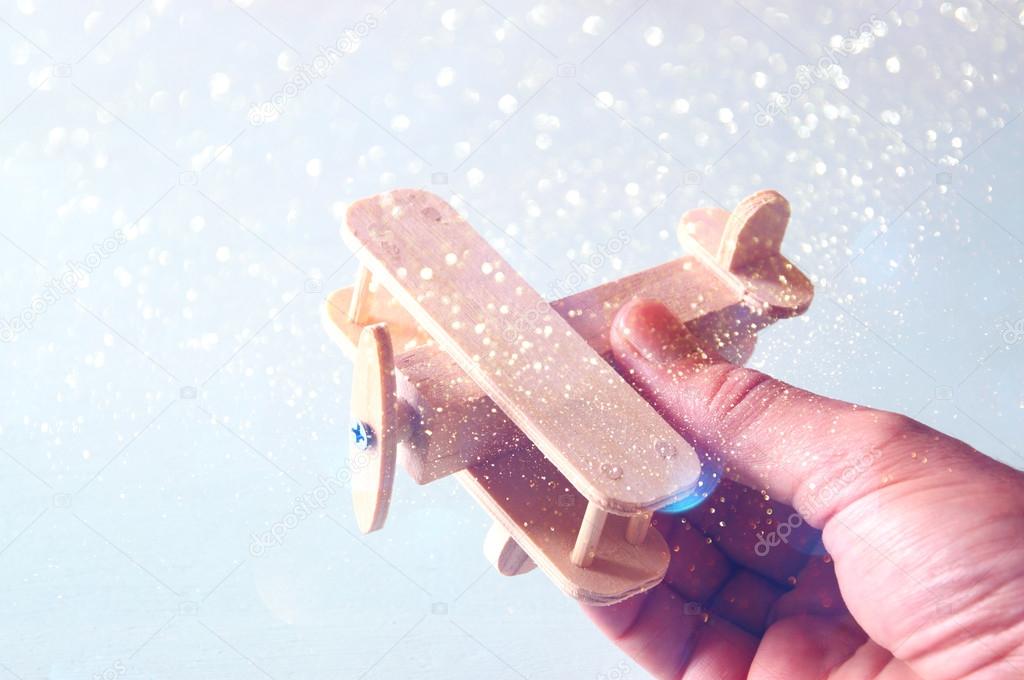 close up photo of man's hand holding wooden toy airplane over wooden background. filtered image with glitter overlay. aspiration and simplicity concept