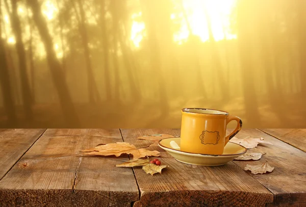 Imagen frontal de la taza de café sobre la mesa de madera y las hojas de otoño frente al fondo otoñal del atardecer . — Foto de Stock