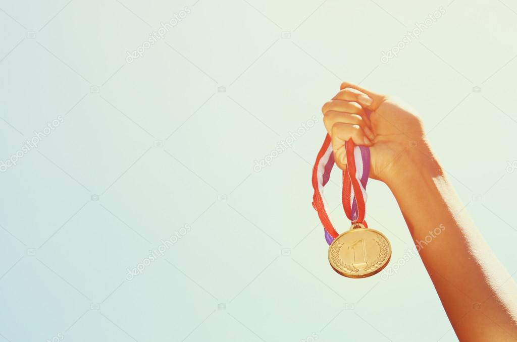 woman hand raised, holding gold medal against sky. award and victory concept.