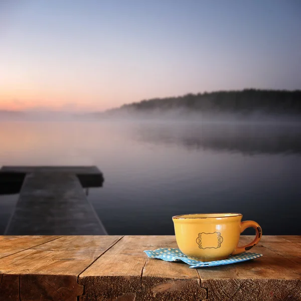 Imagen frontal de la taza de café sobre la mesa de madera frente a la calma niebla vista del lago al atardecer . — Foto de Stock