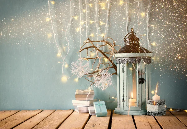 white wooden vintage lantern with burning candle christmas gifts and tree branches on wooden table. retro filtered image with glitter overlay.