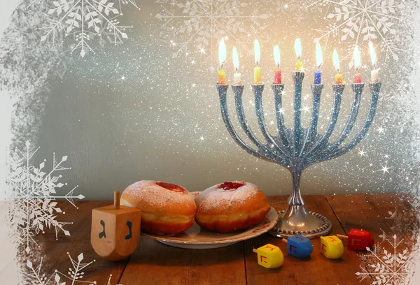 Image of jewish holiday Hanukkah with menorah (traditional Candelabra), donuts and wooden dreidels (spinning top). retro filtered image with glitter and snowflakes overlay. — Stockfoto