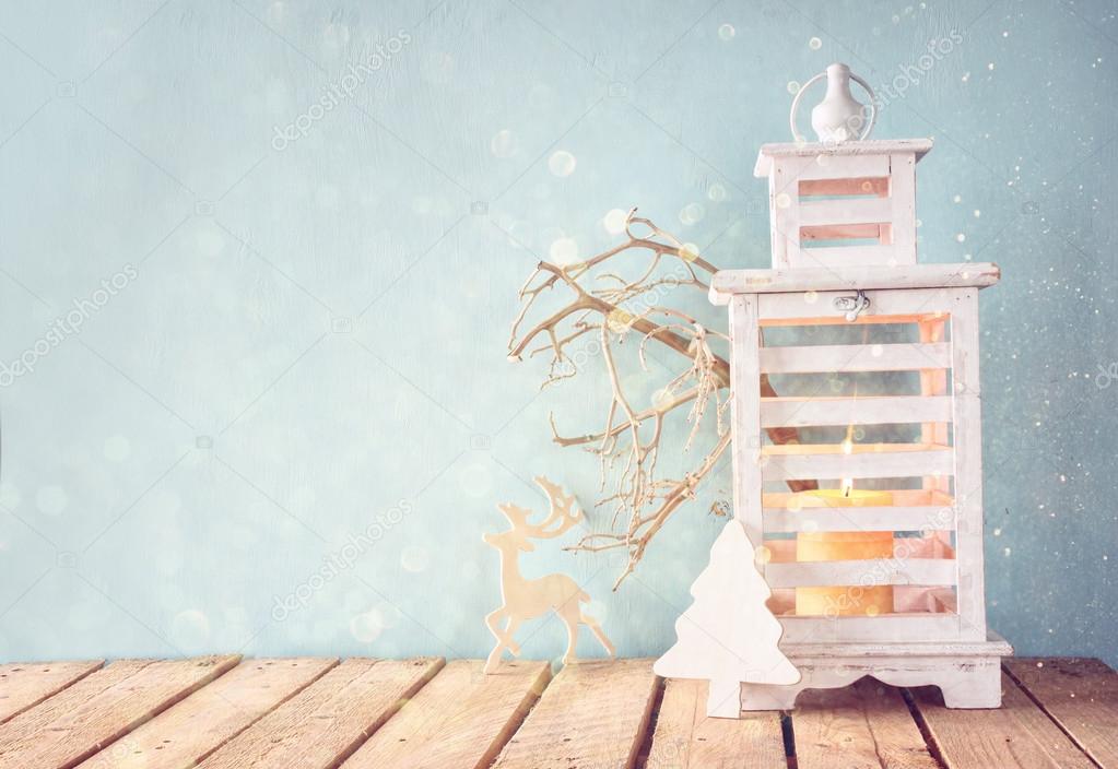white wooden vintage lantern with burning candle, wooden deer, christmas gifts and tree branches on wooden table. retro filtered image with glitter overlay.