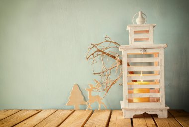 white wooden vintage lantern with burning candle, wooden deer, christmas gifts and tree branches on wooden table. retro filtered.