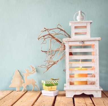 white wooden vintage lantern with burning candle, wooden deer, christmas gifts and tree branches on wooden table. retro filtered image.