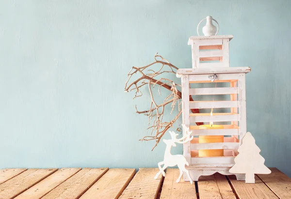 White wooden vintage lantern with burning candle, wooden deer, christmas gifts and tree branches on wooden table. — Stock fotografie