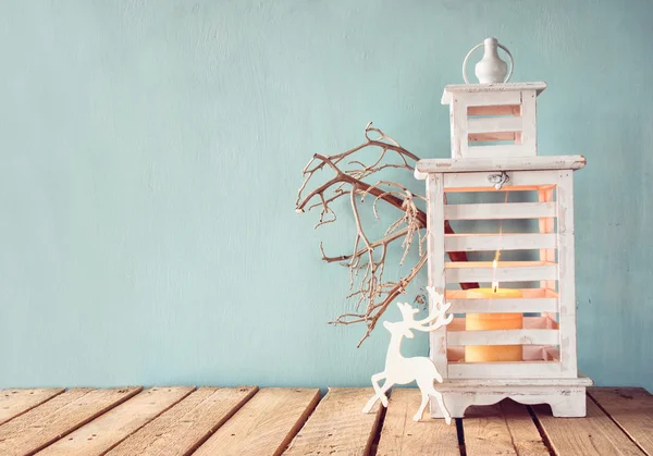 Linterna vintage de madera blanca con vela encendida, ciervos de madera, regalos de Navidad y ramas de árbol en la mesa de madera. retro filtrado . — Foto de Stock