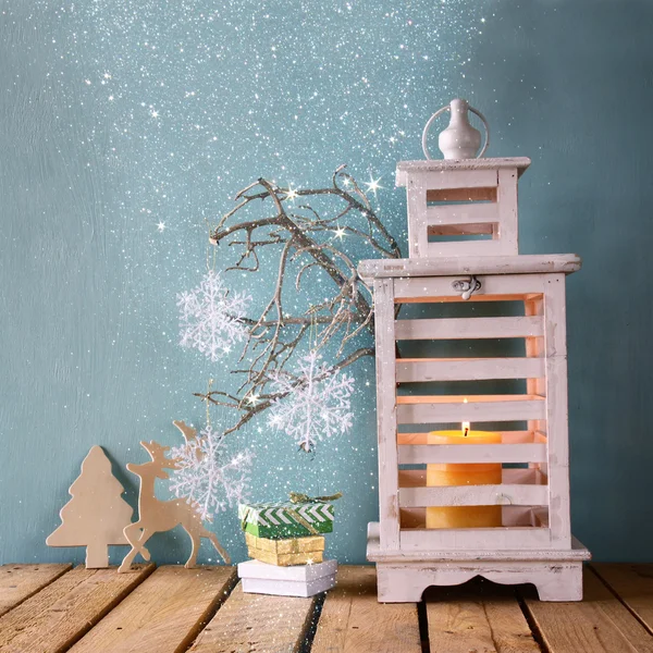White wooden vintage lantern with burning candle, wooden deer, christmas gifts and tree branches on wooden table. retro filtered image with glitter overlay. — Stock Photo, Image