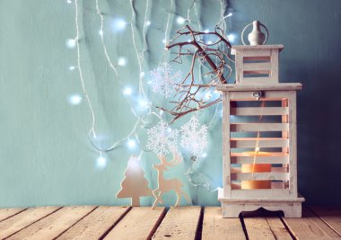 white wooden vintage lantern with burning candle, wooden deer, christmas gifts and tree branches on wooden table. retro filtered image.