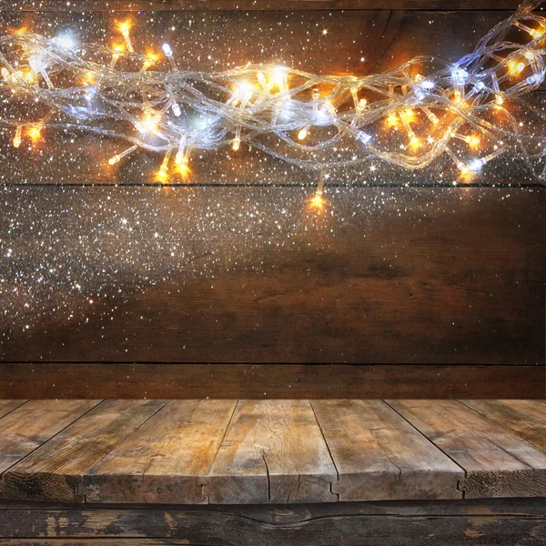 Wood board table in front of Christmas warm gold garland lights on wooden rustic background. filtered image. selective focus. glitter overlay. — Stock Photo, Image