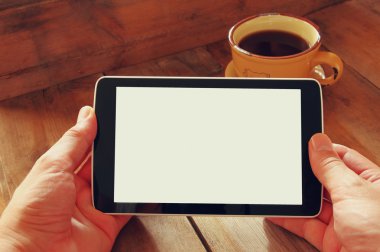 Digital tablet computer with isolated screen in male hands over wooden table background and cup of coffee.