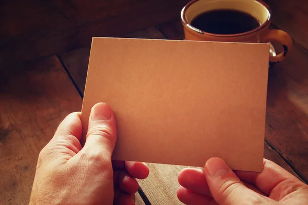 Male hands holding brown empty card over wooden table background and cup of coffee. retro style image, low key and warm tones. — Stock Photo, Image