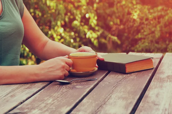 Cerrar imagen de la mujer leyendo libro al aire libre, junto a la mesa de madera y taza de café por la tarde. imagen filtrada. imagen filtrada. enfoque selectivo . — Foto de Stock