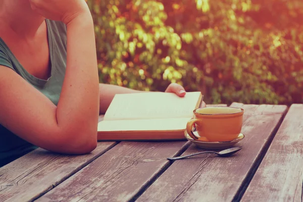 Close up imagem da mulher lendo livro ao ar livre, ao lado de mesa de madeira e xícara de café à tarde. imagem filtrada. imagem filtrada. foco seletivo . — Fotografia de Stock
