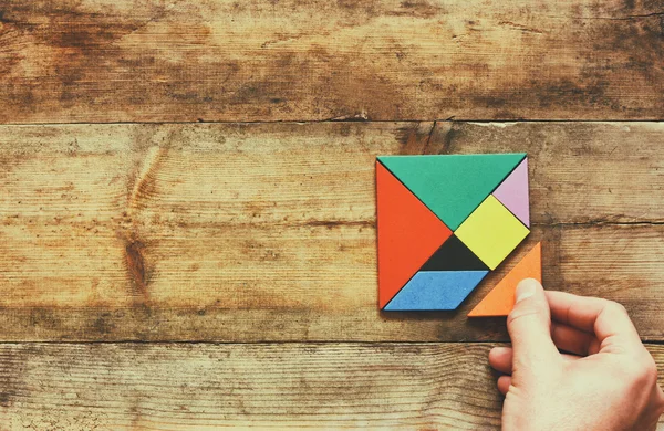 Pohled shora fotografie z lidské ruky držící chybějící kousek v čtvercové tangram puzzle, dřevěný stůl — Stock fotografie