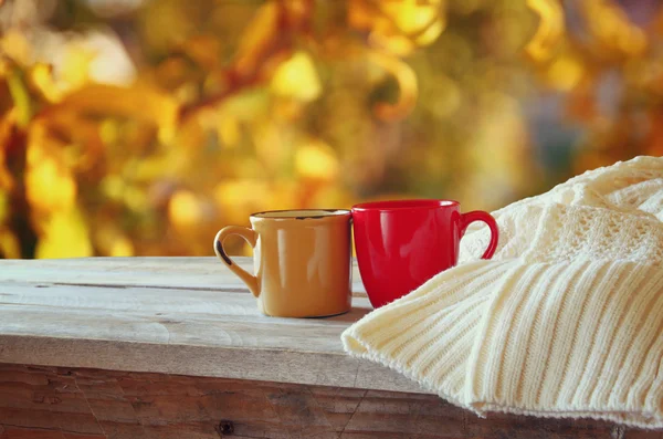 Front image of two coffee cups over wooden table and woolen sweater in front of autumnal sunset background. Valentines Day concept. — Stock Fotó