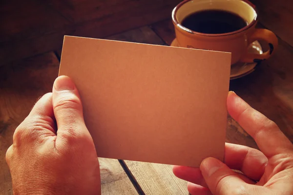 Male hands holding brown empty card over wooden table background and cup of coffee. retro style image, low key and warm tones. — Stock Photo, Image