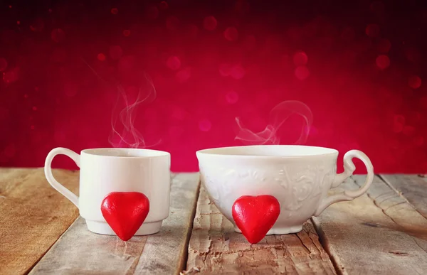 Image of tow red heart shape chocolates and couple cups of coffee on wooden table in front of bokeh abstract background. valentine's day celebration concept. — Zdjęcie stockowe