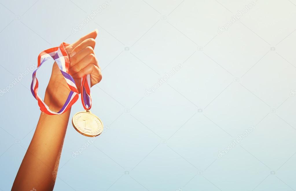 woman hand raised, holding gold medal against sky. award and victory concept.