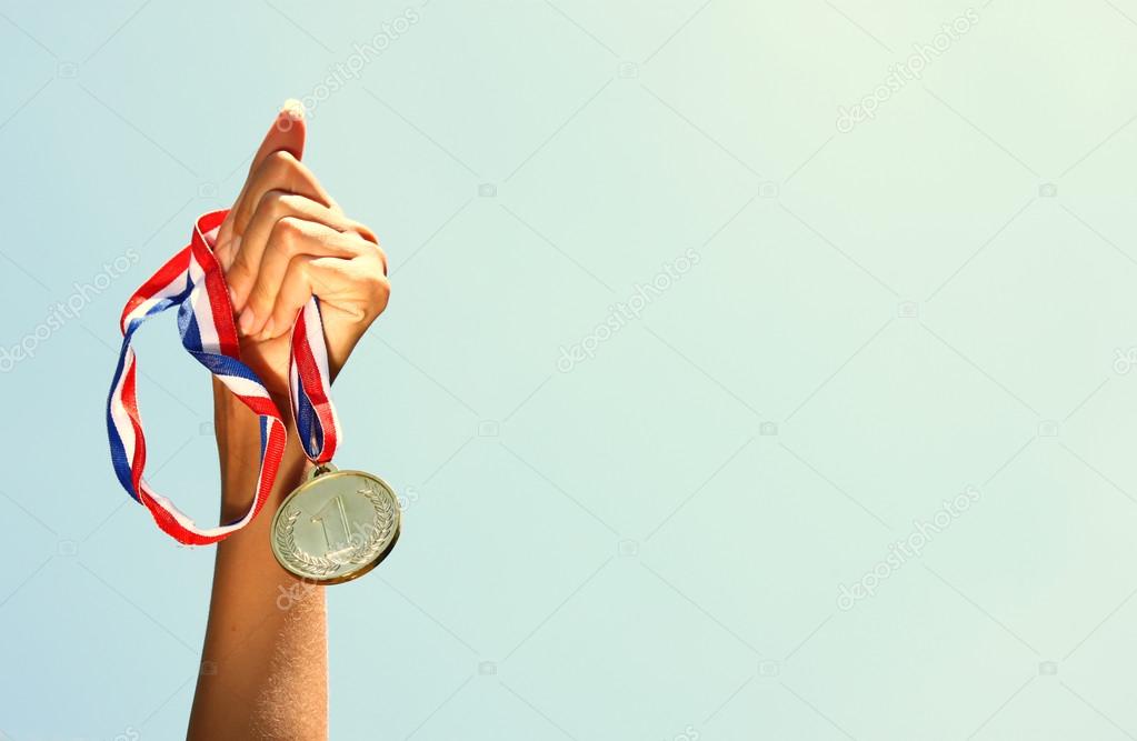 woman hand raised, holding gold medal against sky. award and victory concept.