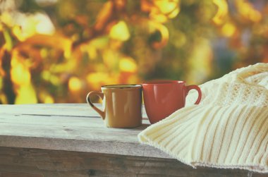 front image of two coffee cups over wooden table and woolen sweater in front of autumnal sunset background. Valentines Day concept.