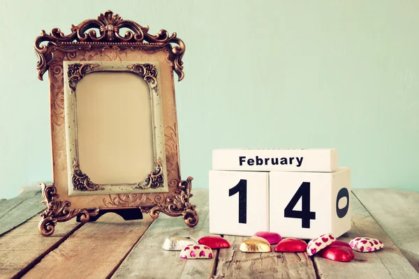 February 14th wooden vintage calendar with colorful heart shape chocolates next to blank vintage frame on wooden table. selective focus.Template ready to put photography