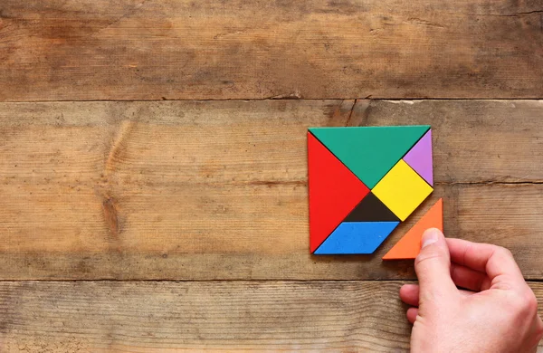 Foto von oben: Männerhand hält ein fehlendes Teil eines quadratischen Tangram-Puzzles über einem Holztisch — Stockfoto