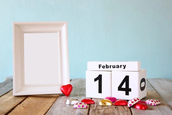 14 февраля деревянный винтажный календарь с красочными шоколадками в форме сердца рядом с пустой винтажной рамой на деревянном столе. Избирательные фокусы. — стоковое фото
