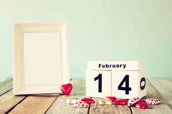 February 14th wooden vintage calendar with colorful heart shape chocolates next to blank vintage frame on wooden table. selective focus.Template ready to put photography. retro filtered — Stockfoto