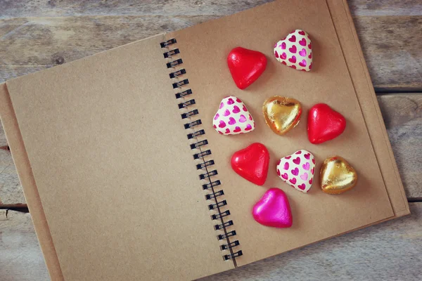 Top view image of colorful heart shape chocolates on open blank notebook on wooden table. valentine's day celebration concept. retro filtered ans toned image — Stockfoto