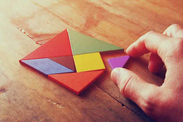 Main d'homme tenant une pièce manquante dans un puzzle carré tangram, sur une table en bois . — Photo