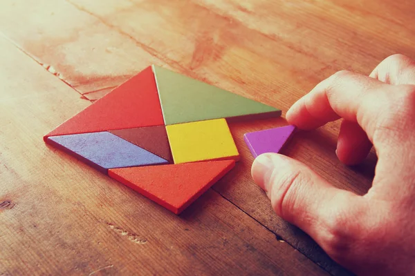 Mano del hombre sosteniendo una pieza perdida en un rompecabezas cuadrado tangram, sobre mesa de madera . — Foto de Stock