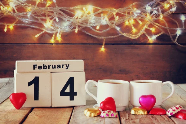 February 14th wooden vintage calendar with colorful heart shape chocolates next to couple cups on wooden table. selective focus. vintage filtered — Stock Photo, Image