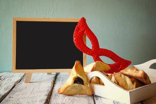 Hamantaschen cookies or hamans ears,noisemaker and mask next to blackboard for Purim celebration (jewish carnival holiday). selective focus — стокове фото